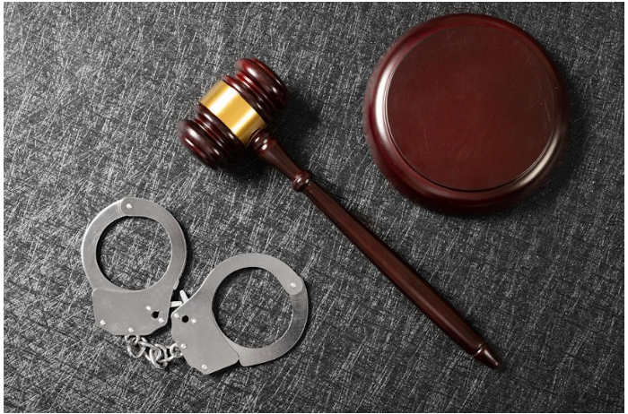 Top-down view of a wooden judge's gavel, sounding block, and steel handcuffs on a dark textured surface, representing services for Criminal DUI Defense Gaylord.
