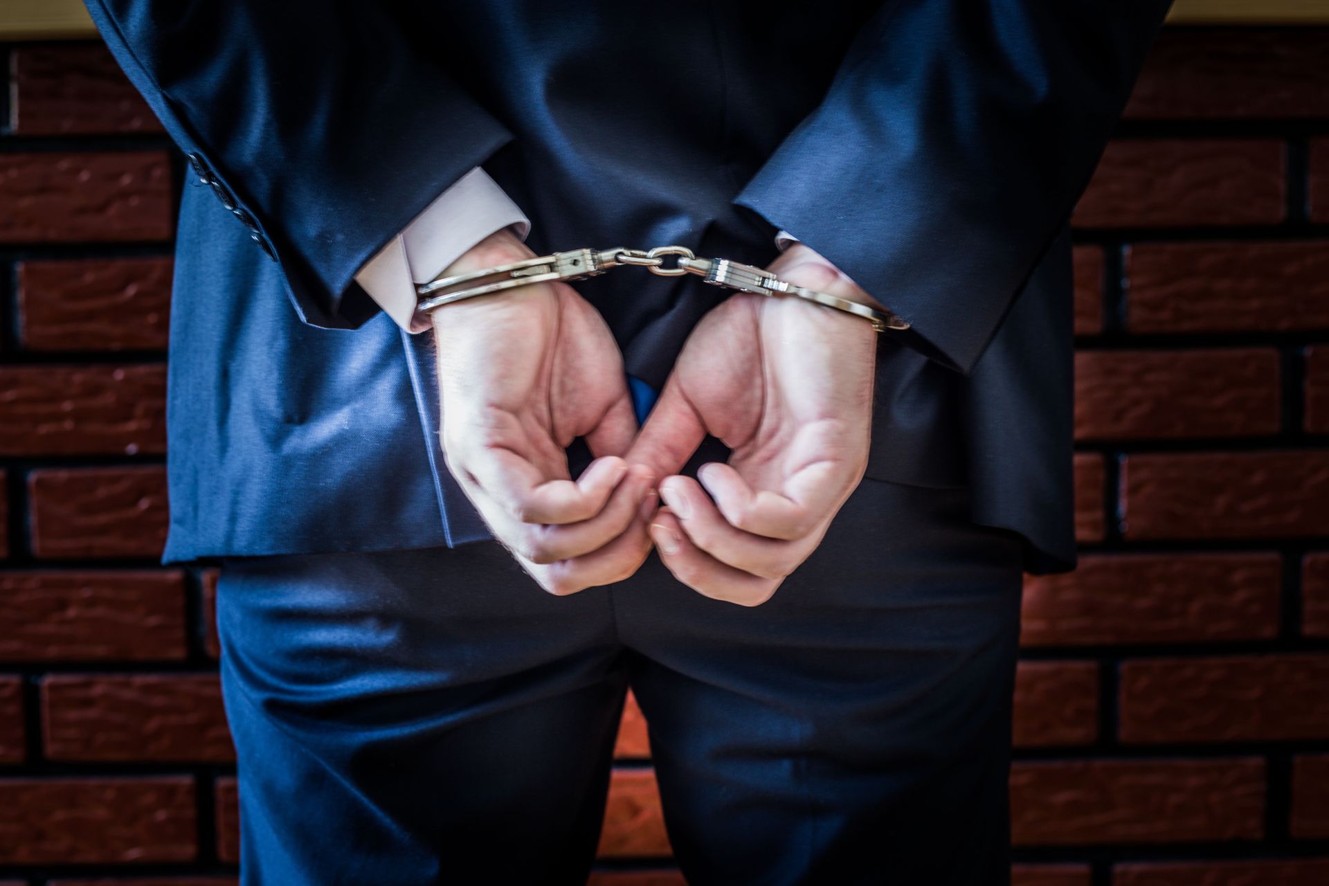 Rear view of a man in a business suit with his hands in handcuffs behind his back, standing against a brick wall, representing an arrest that requires a qualified DUI Criminal Defense Attorney