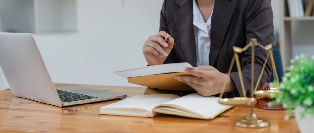 Legal professional in a suit holding case documents at a wooden desk set with a laptop, open book, and scales of justice, working as a DWI Criminal Defense Attorney.